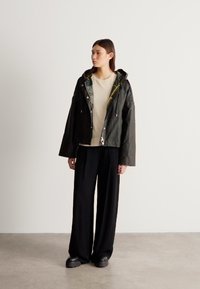 Must kapuutsiga jakk, millel on roheline ja kollane ruuduline vooder, kandmas beige särgi peal. Üheskoos lahtiste mustade pükste ja jämedate mustade kingadega.