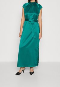 Satin teal maxiklänning med hög halsringning, korta ärmar, knytbara midjeband och en sidoklyvning, tillsammans med svarta spetsiga klackskor.