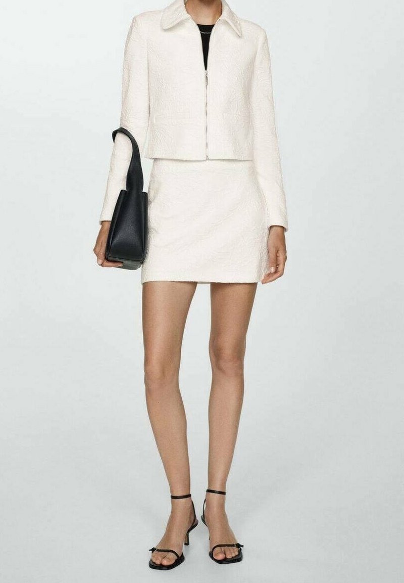 Femme portant un ensemble veste et jupe texturée blanche, tenant un sac à main noir et portant des sandales à talons noires avec bride à la cheville.
