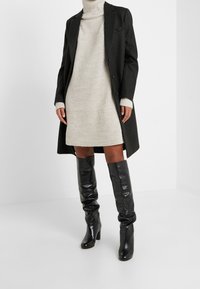 Mujer vistiendo un vestido de punto crema con cuello alto, botas de cuero negras hasta la rodilla y un abrigo negro con botones en un estudio blanco.