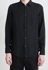 Svart långärmad skjorta i lätt tyg, med knapp-down krage, framficka och blå knappar, samt en något kurvad nederkant.