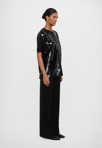 Black sequined top with short sleeves, relaxed fit, paired with wide-leg black pants and flat black shoes. Model stands sideways.