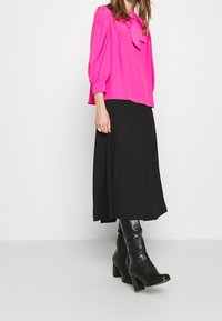 Fuksiapinkki paita, jossa on nauhavyö, yhdistetty mustaan, ilmavaan midi-hameeseen ja polven yli ulottuviin mustiin korkokenkiin. Sujuvat kankaat ja erottuvat muodot.