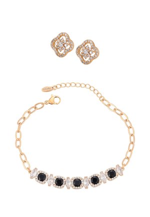 Złota bransoletka z na przemian ułożonymi czarnymi i przezroczystymi kamieniami szlachetnymi, zestawiona ze złotymi kolczykami w kształcie sztyftów ozdobionymi przezroczystymi kamieniami szlachetnymi.
