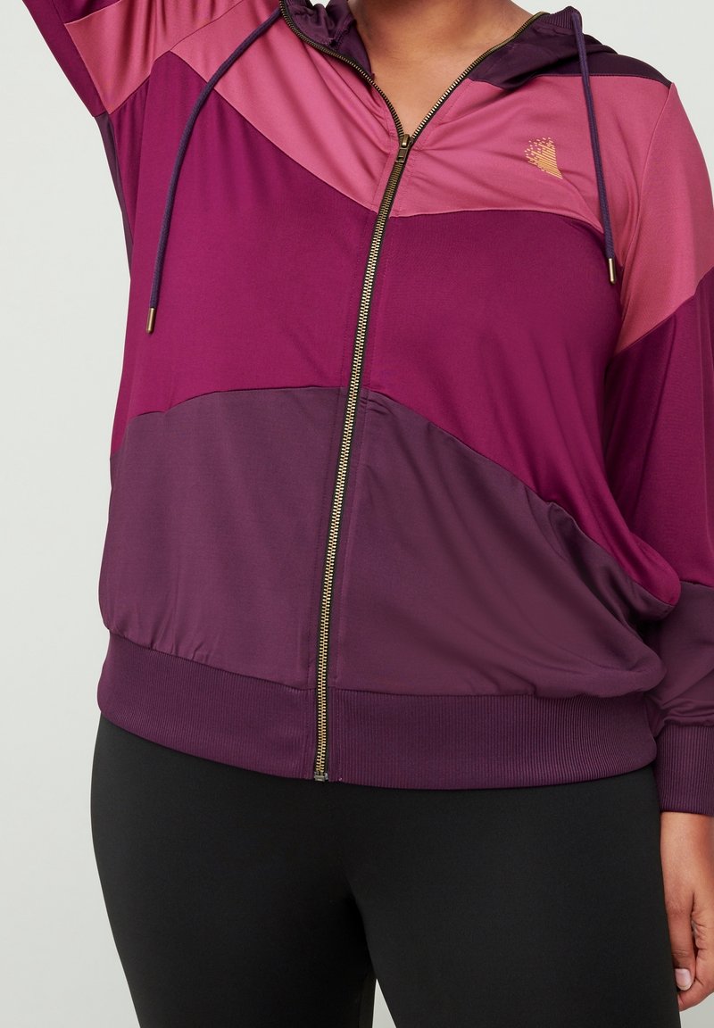 Femme portant un sweat à capuche à fermeture éclair violet à plusieurs nuances avec un petit logo sur la poitrine et un legging noir, bras partiellement levé.