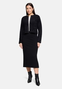 Czarny, fakturowany krótki kardigan z guzikami, w zestawie z dopasowaną czarną midi spódnicą. Modelka nosi czarne, spiczaste botki za kostkę. Pod spodem neutralna bluzka.