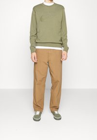 Sweatshirt Nike vert olive avec logo en relief, associé à un pantalon marron clair et des baskets vertes, avec des lacets et des accents blancs.