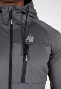 Grijze sportieve hoodie met rits aan de voorkant, voorzien van een klein logo op de borst, gestructureerd materiaal, en verstelbare capuchon met trekkoorden.