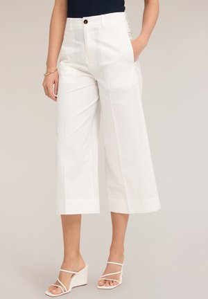 Femme portant un pantalon blanc large et court, un top noir sans manches, des sandales blanches compensées à brides et un bracelet doré au poignet gauche.