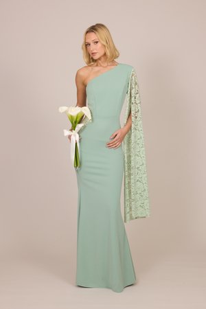 Vrouw in een lichtgroene een-schouderjurk met kanten mouw houdt witte calla's vast, gebonden met een wit lint tegen een effen beige achtergrond.