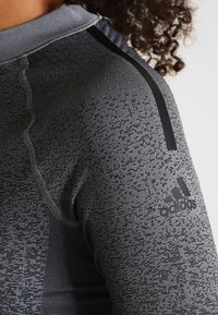 Szara koszulka sportowa z teksturowanej tkaniny, kontrastowymi czarnymi akcentami oraz logo Adidas na rękawie. Zbliżenie na strefę ramienia.