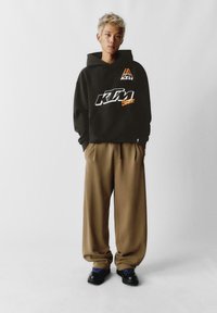 Čierna mikina s kapucňou s logom KTM v bielej a oranžovej farbe, v kombinácii s hnedými širokými nohavicami a čiernymi topánkami s modrými šnúrkami.