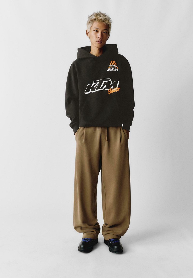 Čierna mikina s kapucňou s logom KTM v bielej a oranžovej farbe, v kombinácii s hnedými širokými nohavicami a čiernymi topánkami s modrými šnúrkami.