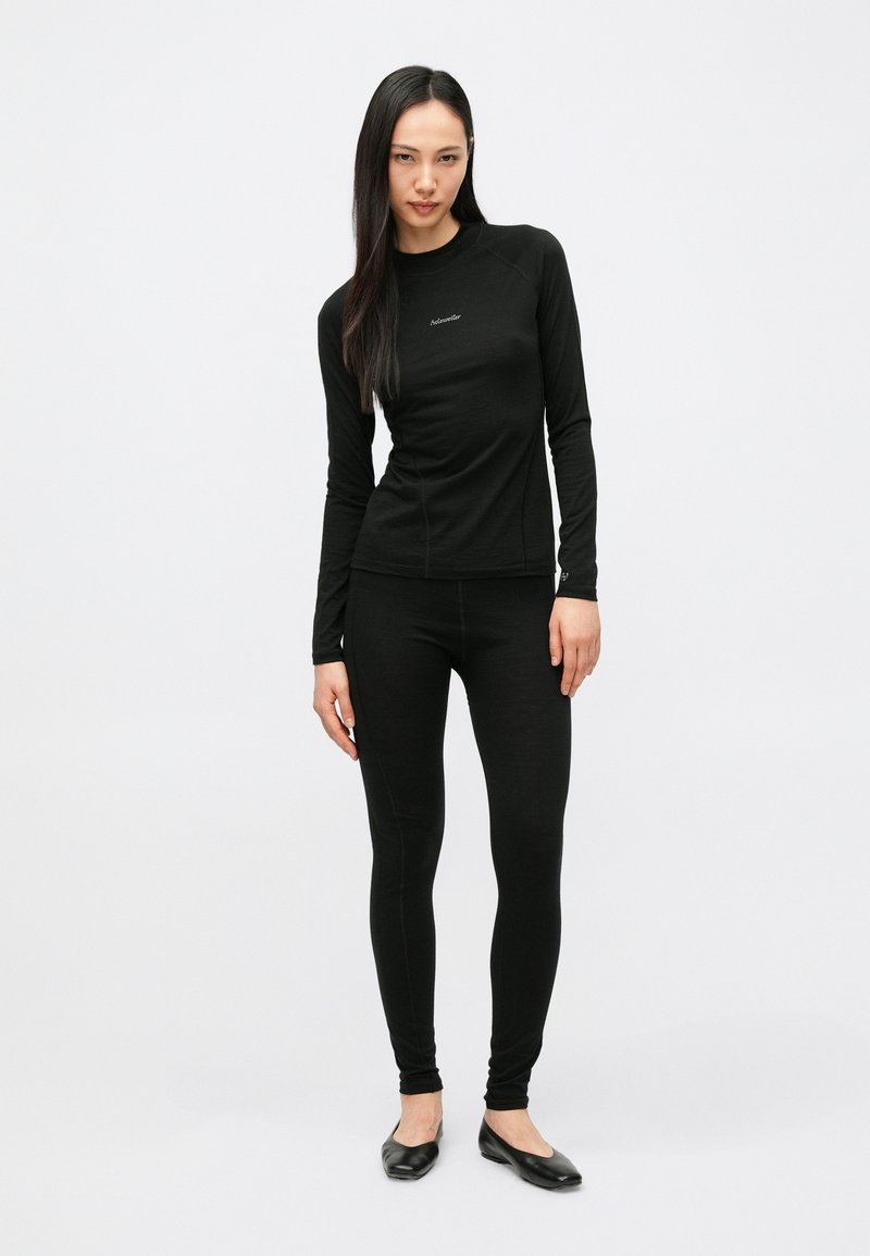 Woman standing against white background wearing fitted black long-sleeve top and leggings with black flats, looking directly ahead.