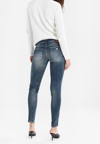Jeans ajustados en denim azul oscuro con un lavado desgastado, que presentan una cintura de altura media y dos bolsillos traseros. Combinados con una blusa blanca y tacones negros.