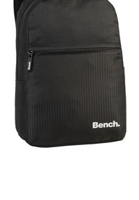 Schwarzer Rucksack mit einem strukturierten hexagonalen Muster im unteren Bereich, einer vorderen Reißverschlusstasche und dem weißen "Bench."-Logo an der Seite.