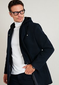 Chaqueta negra con capucha, con cremallera frontal, dos bolsillos laterales y una textura suave, combinada con un cuello alto blanco debajo.