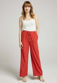 Pantalons larges, légers, rouges avec une taille élastique et un cordon de serrage, associés à un débardeur blanc ajusté et des chaussures beiges.