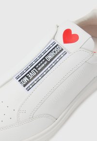 Balti odiniai sportbačiai, kuriuose yra juoda ir balta etiketė su tekstu, raudonas širdelės akcentas ir įspaustas "LOVE MOSCHINO" šone.