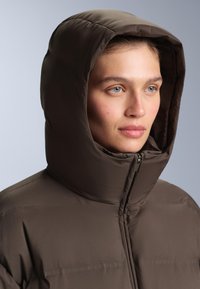 Hnedá zamknutá bunda s vysokým golierom, plným zipsovým uzáverom a veľkou kapucňou. Hladký povrch s prešitými detailmi na látkovom povrchu.