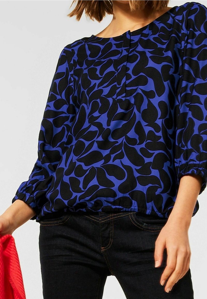 Femme portant un chemisier bleu avec des formes noires abstraites et un jean foncé, tenant un tissu rouge dans une main sur un fond uni.