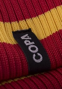 Rood en geel gebreid textiel met een gestructureerd oppervlak; zwart label met "Copa" in witte tekst bevestigd aan de rand.