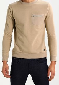 Felpa beige a maniche lunghe con scollo rotondo, tessuto strutturato, tasca sul petto con cerniera e polsini e orlo a costine.