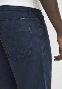 Navy blue denim pants with a straight cut, featuring a back pocket, visible stitching, and a tag that reads "DESIGNED TO DENIM PERFECTION."