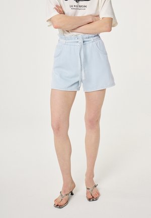 Persoon draagt een wit T-shirt met grafische print, lichtblauwe paperbag shorts met riem en open sandalen met hoge hak en slang-print.