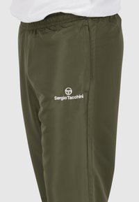 Olijfgroene sportbroek van lichtgewicht materiaal, met een elastische tailleband en een wit logo aan de linkerkant.