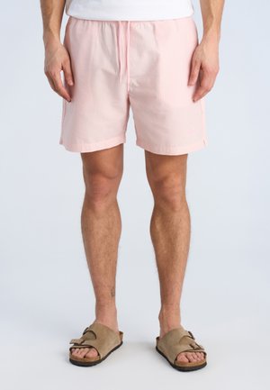 Mand iført lyserøde teksturerede shorts med snøre og beige slip-on sandaler, stående mod en ensfarvet baggrund.