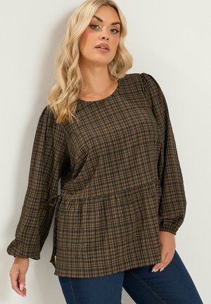 Kobieta o długich blond włosach nosi brązową i czarną koszulę w kratę z długim rękawem i marszczoną talią oraz niebieskie dżinsy, stoi oparta o białą ścianę.