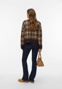 Sweter z wełny w kratę w odcieniach brązu i beżu, krótki fason z długimi rękawami. Ciemne dzwony z denimu, beżowe obuwie i mała brązowa torebka.