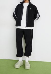 Zwarte fleece zip-up jas met witte strepen en logo, gecombineerd met bijpassende broek. Lichtgroene klompen en witte kousen maken de look compleet.