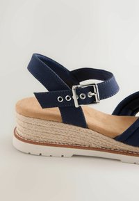 Sandale compensée bleu marine avec bride réglable à boucle argentée, semelle en daim marron clair, plateforme enveloppée de jute et semelle en caoutchouc blanche.