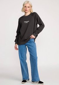 Dunkelgrauer Sweatshirt mit weißem Logo, kombiniert mit hellblauen Schlagjeans und schwarz-weißen Sneakers. Mit Raglanärmeln.