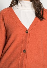 Orange ullcardigan med V-ringning, med knäppning och mjuk textur, över en krämfärgad ribbad polotröja.