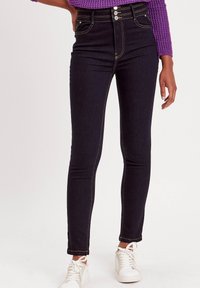 Jean skinny noir taille haute en denim avec des coutures dorées. Comprend une fermeture à trois boutons, cinq poches et une coupe ajustée le long des jambes.