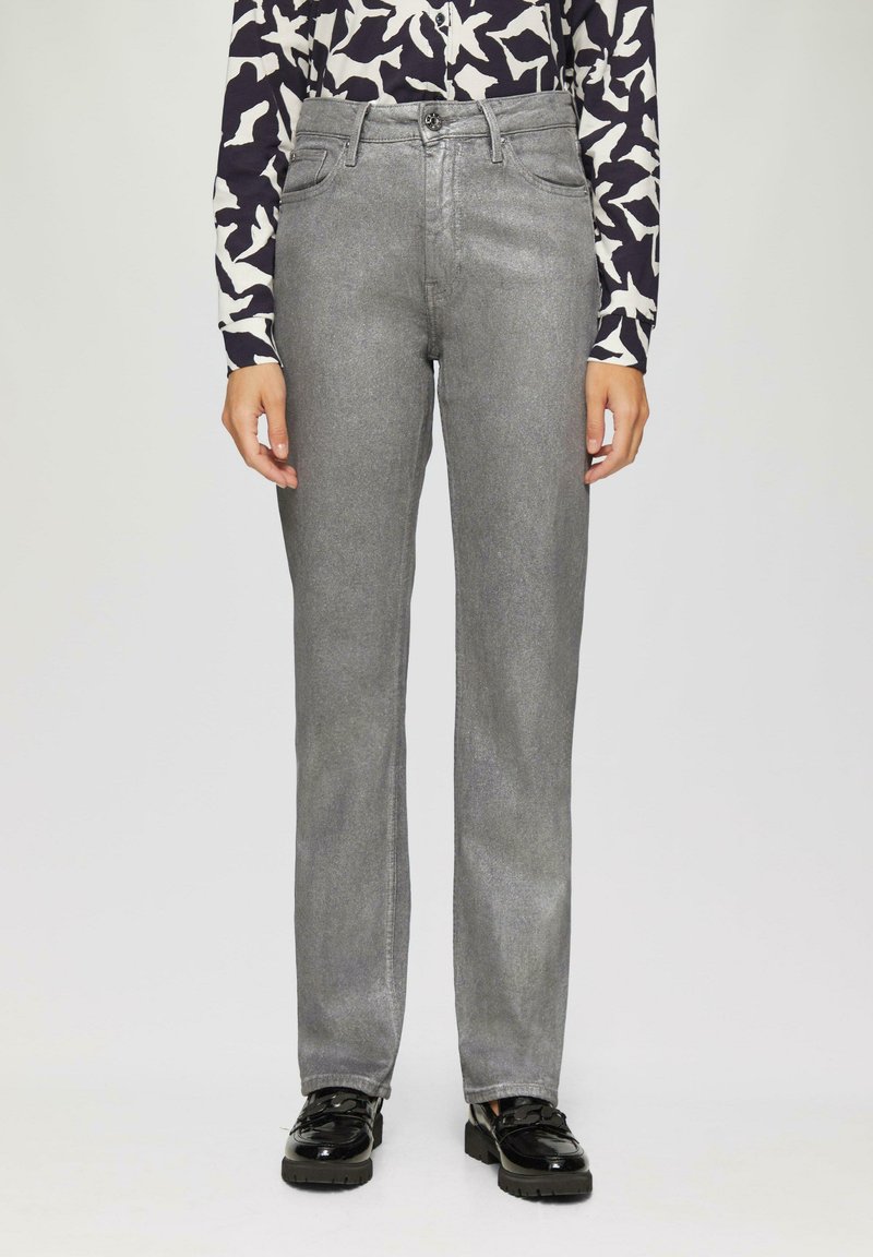 Jean en denim gris avec une coupe droite, présentant une fermeture à bouton et des passants de ceinture. Associé à une chemise à manches longues à motifs et des chaussures noires.