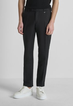 Persona con pantalones negros a medida y zapatillas blancas de pie frente a un fondo gris liso.