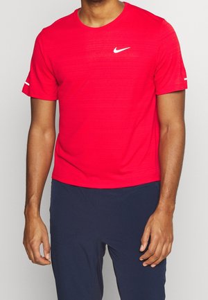 Mann trägt ein leuchtend rotes Nike-Sportshirt mit kurzen Ärmeln und dunkelblaue Hose, steht vor einem neutralen Hintergrund.