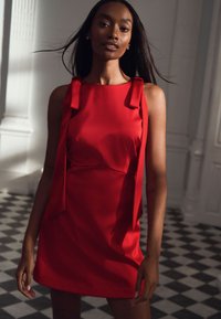 Raudona be rankovių suknelė su dekoratyviais raišiais ant pečių, pagaminta iš lygaus audinio, pasižyminti prigludusiu siluetu ir glotnia tekstūra.