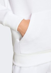 Biely sveter s dlhým rukávom, s rebrovanými manžetami a prednou vreckom. Hladká textúra s voľným strihom a viditeľnými švami.