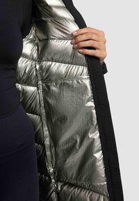 Piumino nero con interno metallico argento, caratterizzato da sezioni trapuntate e un tessuto esterno liscio. Mano che afferra l'apertura.