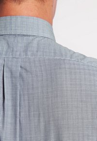 Bakåtsidan av en person som bär en ljusgrå rutad skjorta med knäppt krage, vilket visar tygmönstret och texturen.