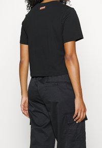Cropped black cotton t-shirt with short sleeves, featuring a small red and white striped tag on the back. Worn with black cargo pants.