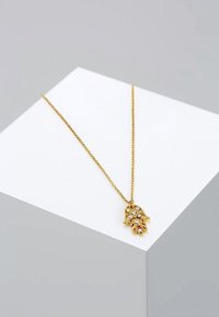 Collier chaîne en or avec un pendentif main de Hamsa en or décoré de petites pierres précieuses claires, présenté sur une surface blanche inclinée.