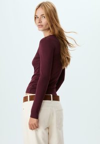 Burgundy long-sleeve top with a fitted shape, paired with cream-colored loose pants and a brown belt. Smooth fabric with minimal texture.