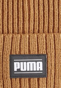 Bonnet en tricot brun avec des côtes verticales. Présente une étiquette rectangulaire noire avec le logo "PUMA" en blanc et des lignes en accent argenté.
