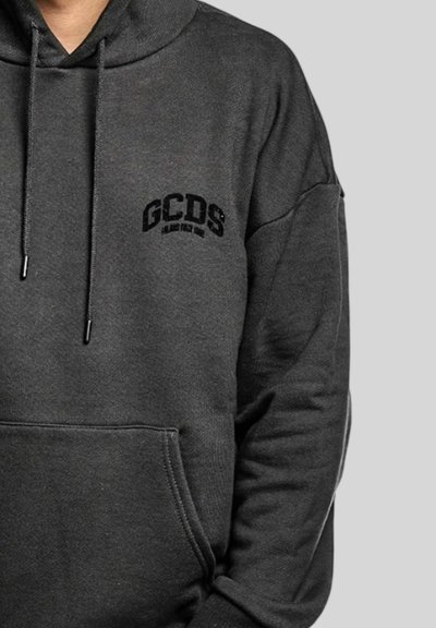 Sweat à capuche gris foncé avec poche avant et logo "GCDS" noir sur le côté gauche de la poitrine, porté par une personne devant un fond gris clair uni.